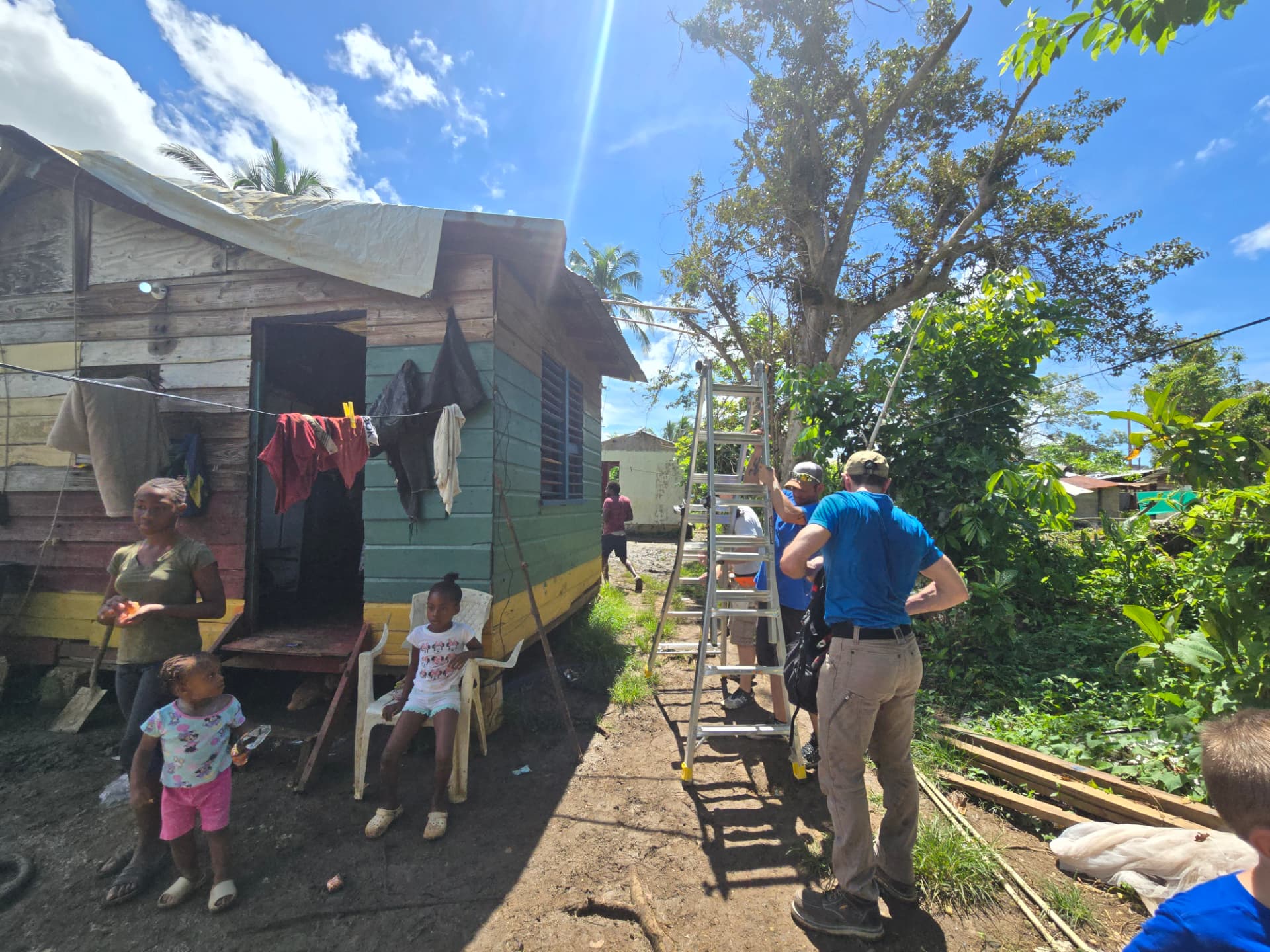 Relief volunteers setting a ladder to repair a tarped roof while a mother and her children stand outside their home