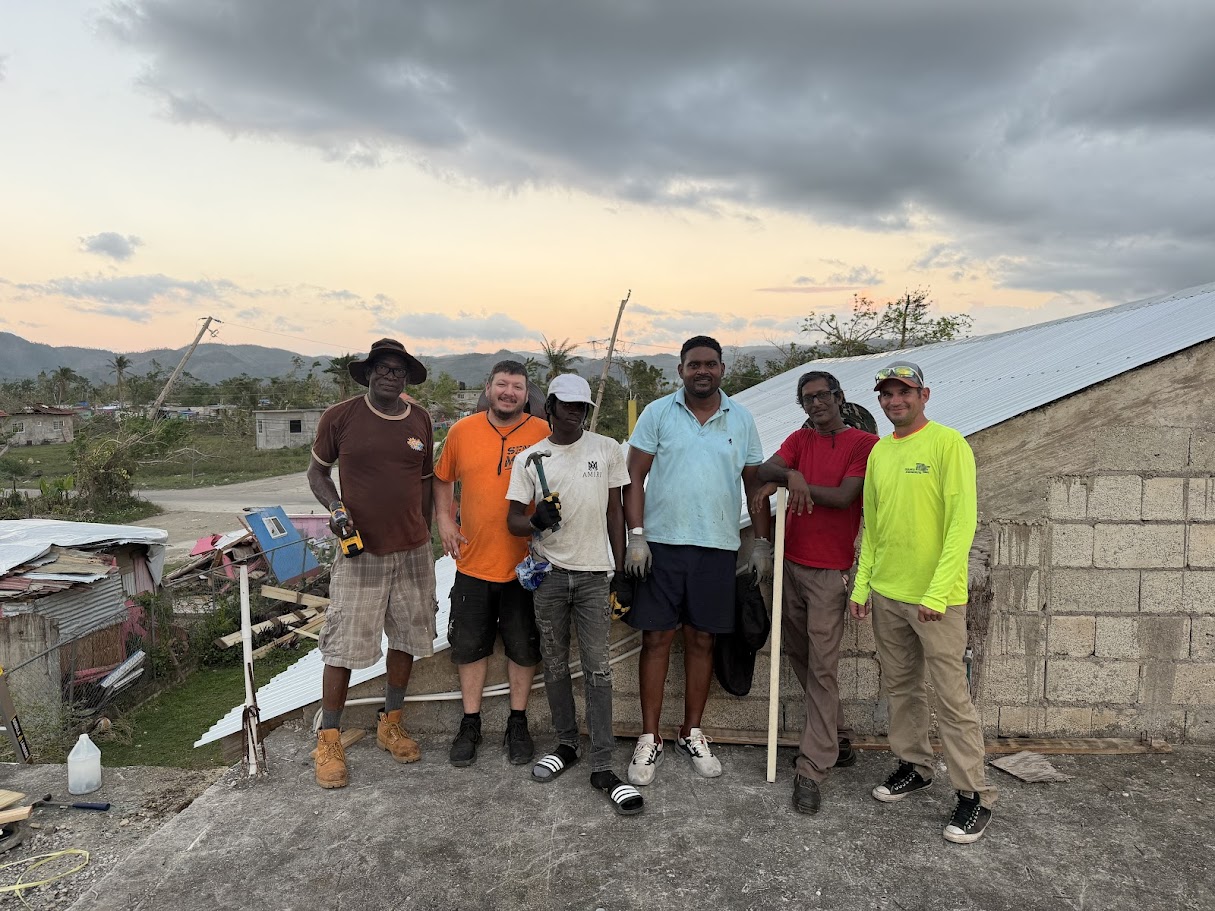 Volunteers and local workers standing together on a rooftop rebuilding site in Jamaica at sunset